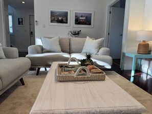 A living room with a beige sofa, a coffee table with a woven tray, and framed pictures on the wall. - Crab Shack Cottage: 4BR in Russell with Pool & Spa, 300m to Waterfront (Russell)