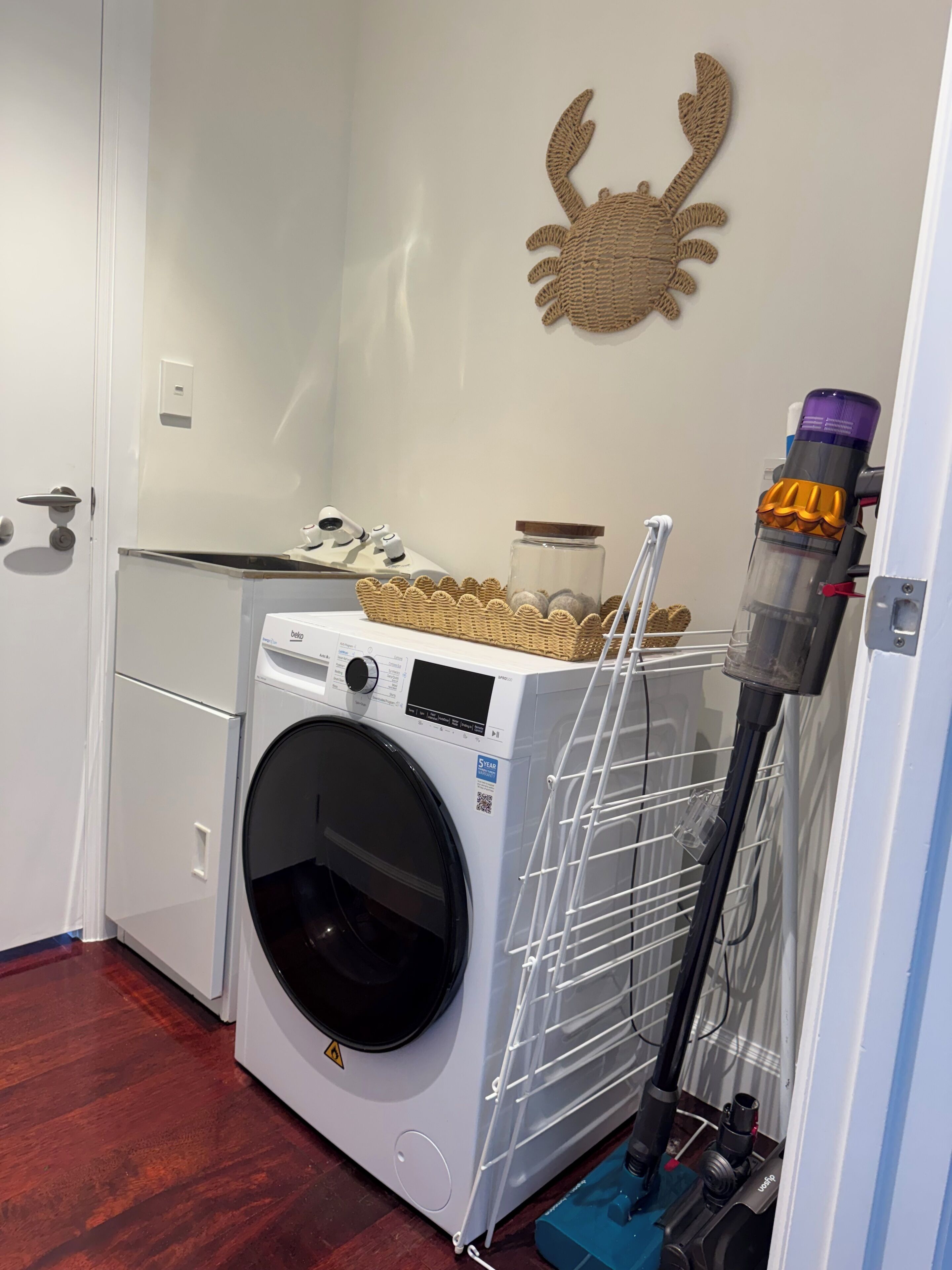 A compact laundry room with a front-loading washing machine, a drying rack, and a vacuum cleaner.