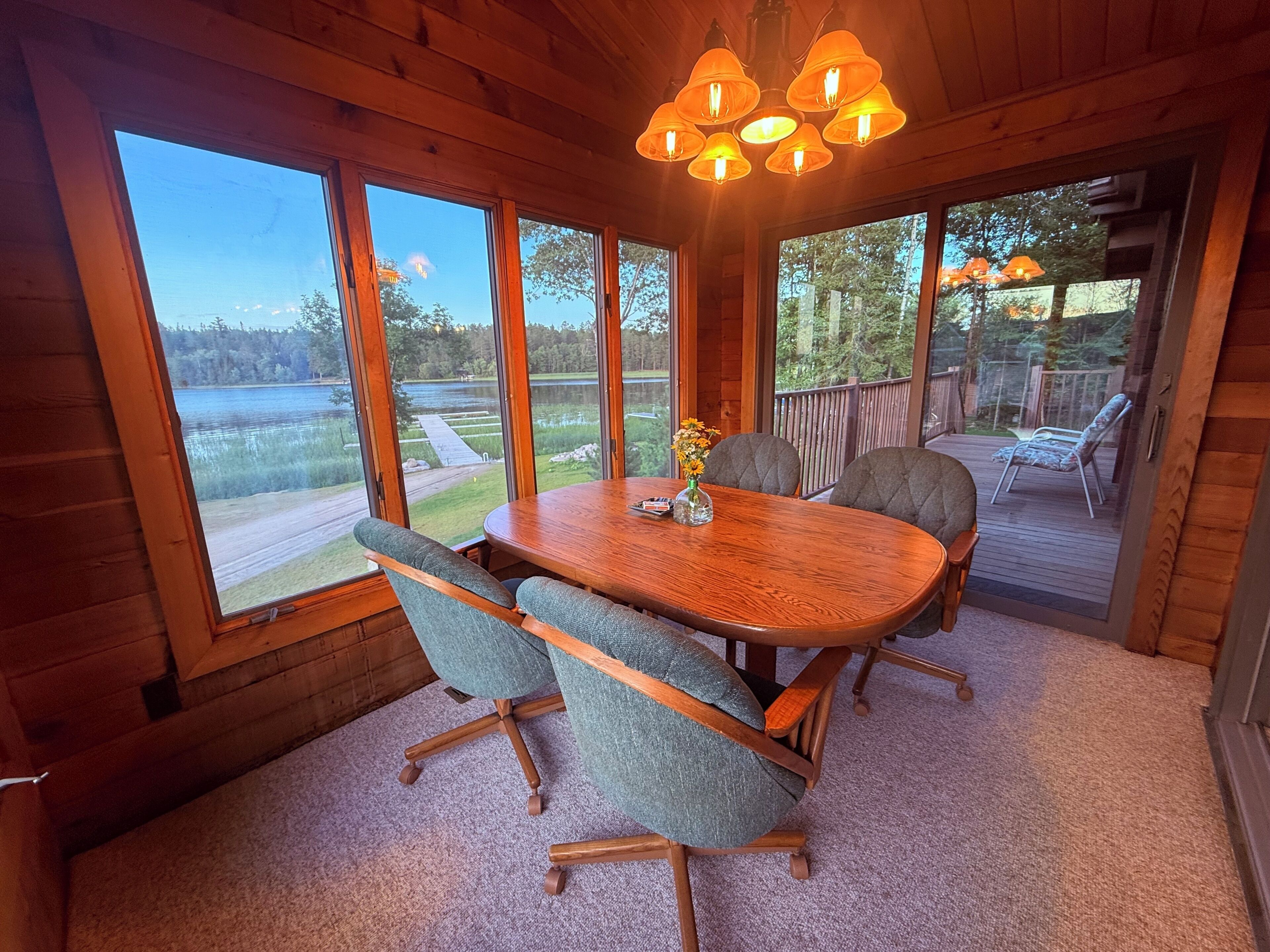 A wooden interior with a dining table, chairs, and large windows overlooking a lake.