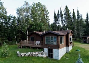 A cabin with a deck, surrounded by a stone wall and a grassy yard.