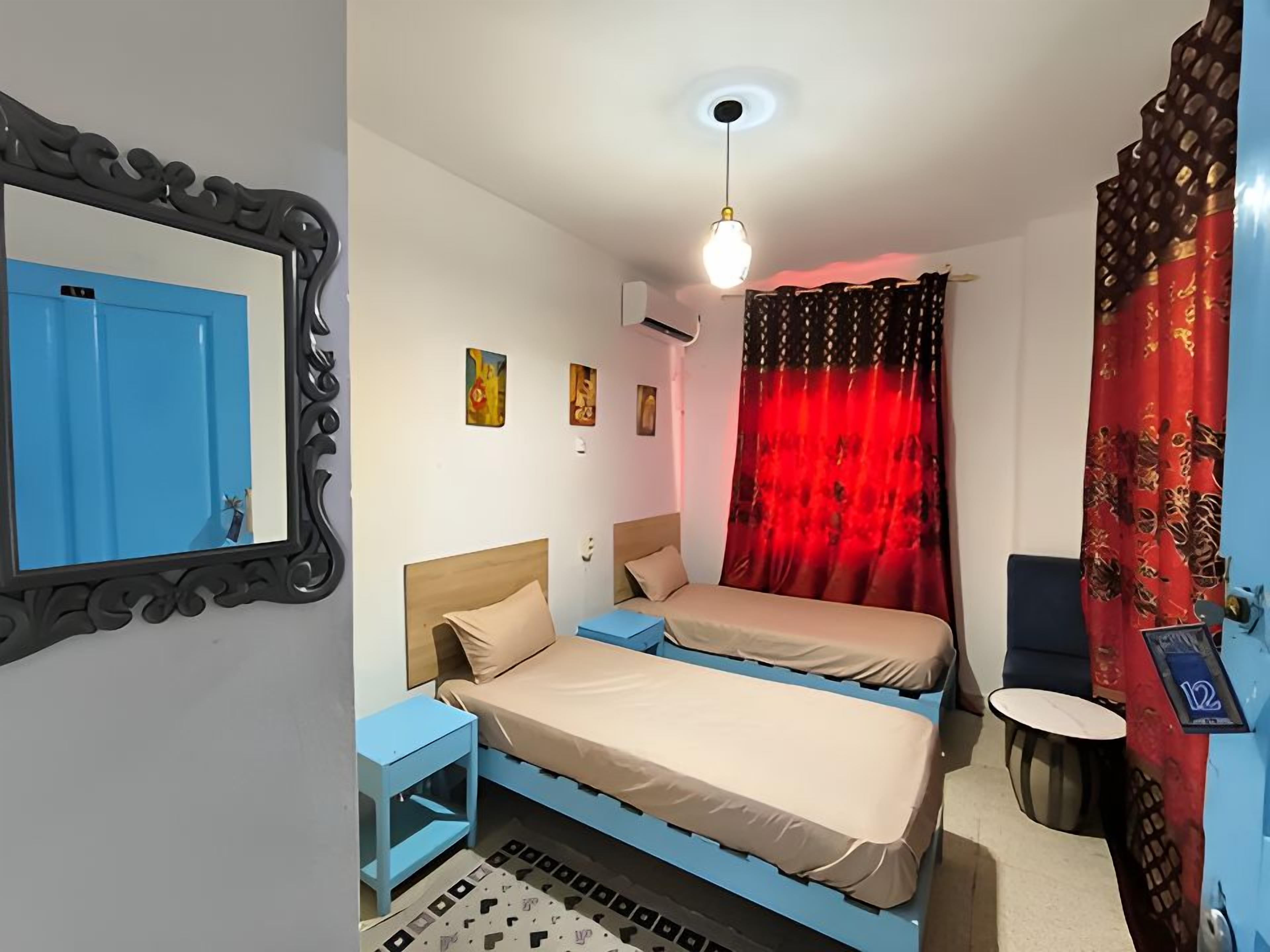 Una habitación con dos camas, una pared azul, una cortina roja, una mesa redonda y una lámpara en el techo.