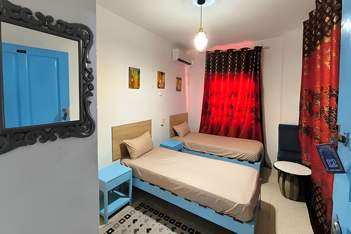A room with two beds, a blue wall, a red curtain, a round table, and a ceiling light.