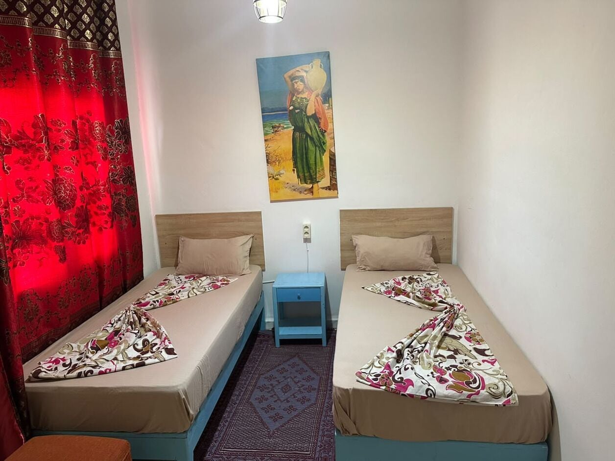 Een kamer met twee eenpersoonsbedden, een nachtkastje, een rood gordijn en een schilderij aan de muur.