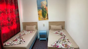 Ein Zimmer mit zwei Einzelbetten, einem Nachttisch, einem roten Vorhang und einem Bild an der Wand.