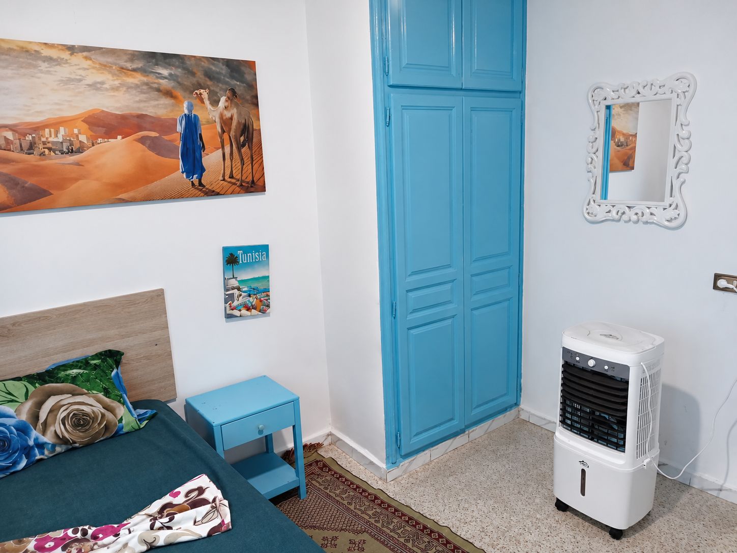 Un dormitorio con una cama, una mesita de noche azul, un calentador de pared, un cuadro enmarcado y un cartel.