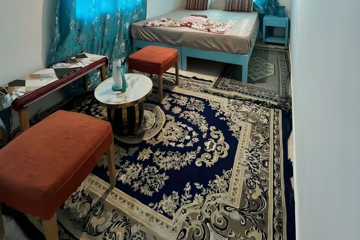 A compact room with a bed, a small table, and a chair. There is a window with curtains, a hanging light fixture, and a patterned rug on the floor.