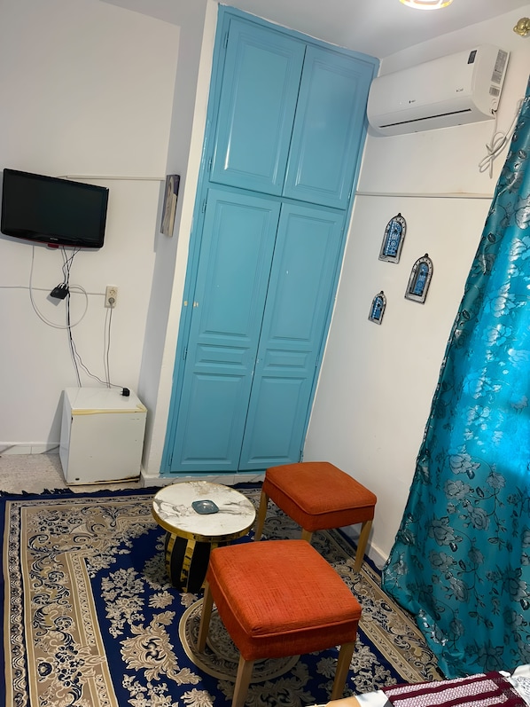 Una habitación con un armario azul, un televisor montado en la pared, una mesita con un objeto decorativo y una alfombra estampada.