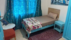 A single-bed room with a patterned bedspread, a small bedside table, a chair, and a painting on the wall.