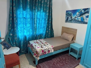 A single-bed room with a patterned bedspread, a small bedside table, a chair, and a painting on the wall.