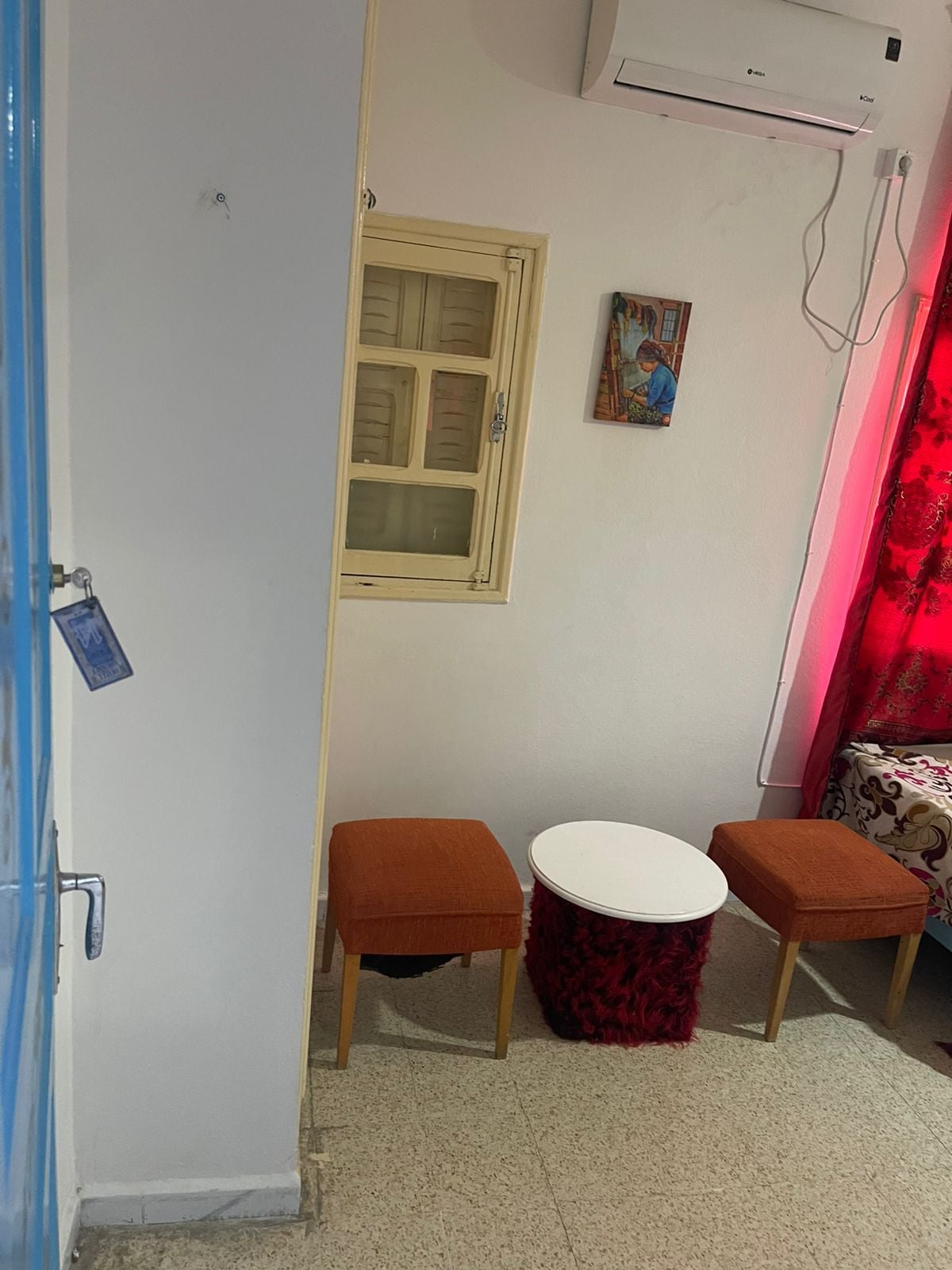 Una habitación pequeña con pared blanca, ventana con marco de madera, cortina roja, mesa redonda pequeña con dos taburetes a juego y un equipo de aire acondicionado montado en la pared.