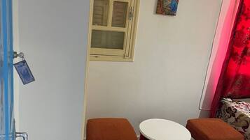 Una habitación pequeña con pared blanca, ventana con marco de madera, cortina roja, mesa redonda pequeña con dos taburetes a juego y un equipo de aire acondicionado montado en la pared.