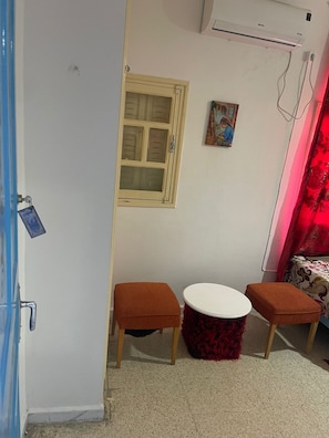 Quarto pequeno com parede branca, janela com moldura de madeira, cortina vermelha, mesa redonda pequena com dois banquinhos combinando e um aparelho de ar condicionado fixado na parede.