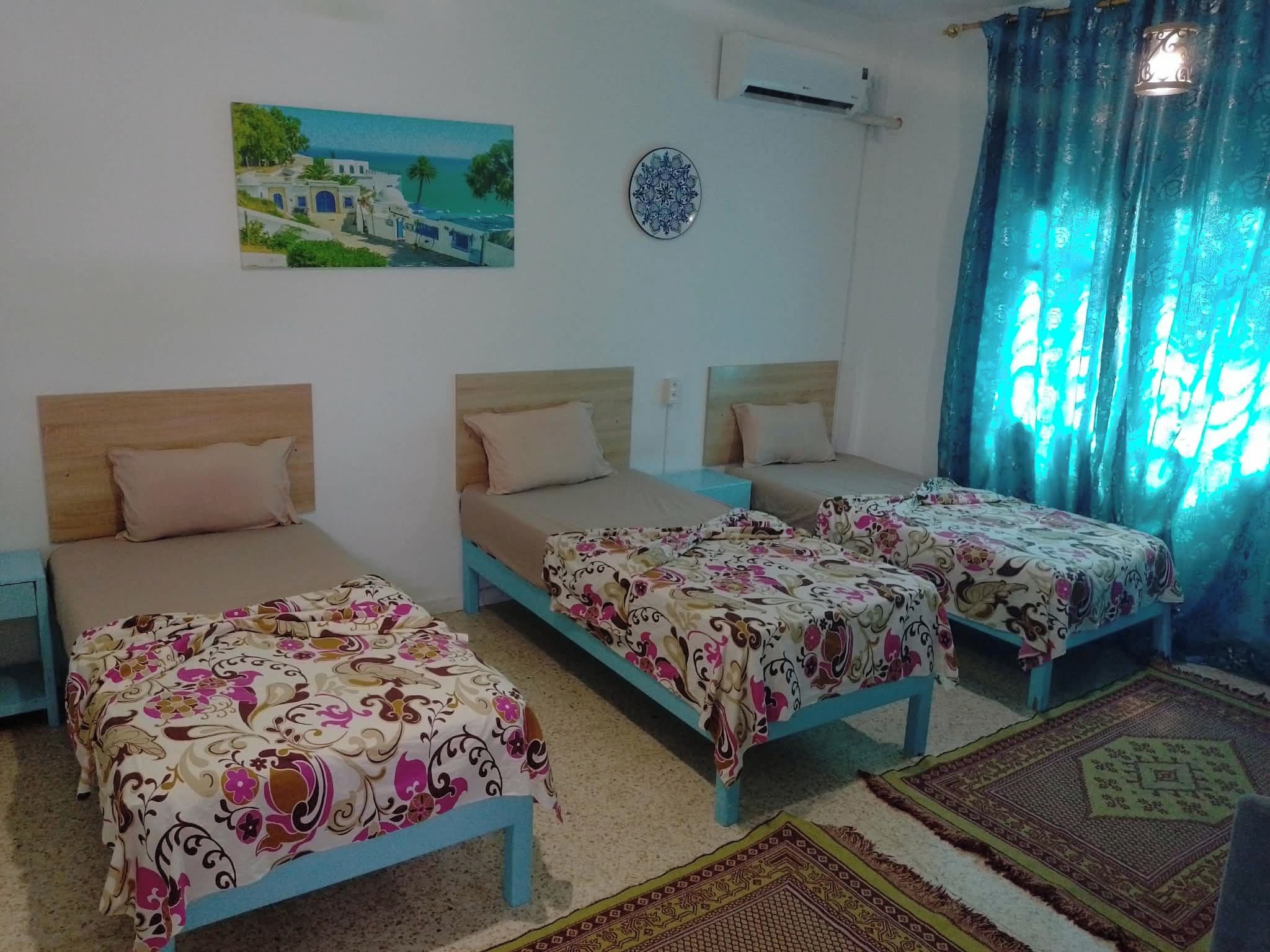 Habitación de hotel con tres camas, un cuadro de una escena costera, un equipo de aire acondicionado y una alfombra estampada.