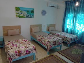 Quarto de hotel com três camas, um quadro de uma paisagem costeira, um aparelho de ar condicionado e um tapete estampado.