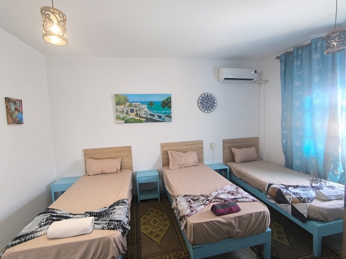 A room with three single beds, a ceiling light, an air conditioner, and a window with curtains.