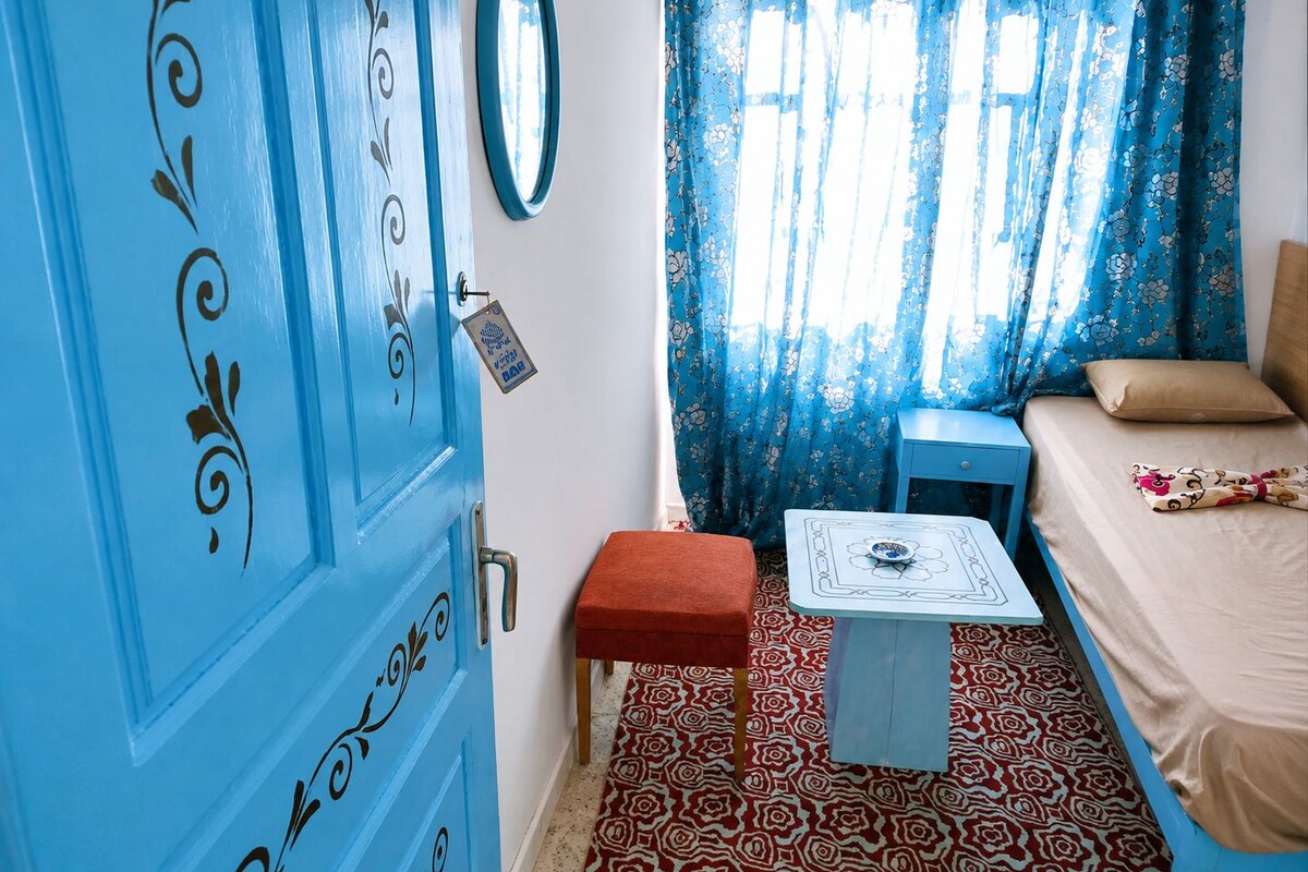 A room with a blue door, a bed with a patterned bedspread, a small table with a decorative item, and a patterned carpet.