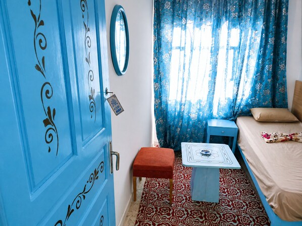 Une pièce avec une porte bleue, un lit recouvert d’une couvre-lit à motifs, une petite table ornée d’un objet décoratif et un tapis à motifs.