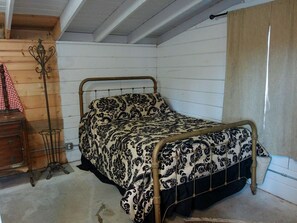 Una cama individual con estructura metálica y ropa de cama estampada en una habitación con paredes blancas y techo de madera.