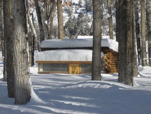 Eine Holzhütte mit schneebedecktem Dach, umgeben von Bäumen in einer verschneiten Landschaft.