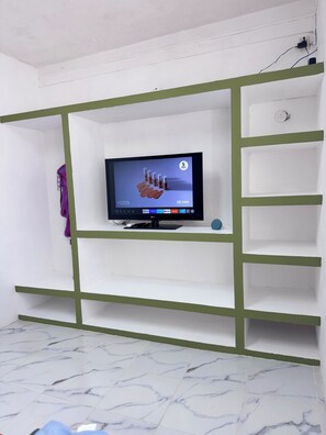 Una habitación con una televisión sobre un estante blanco, un objeto morado a la izquierda y un suelo con estampado de mármol.