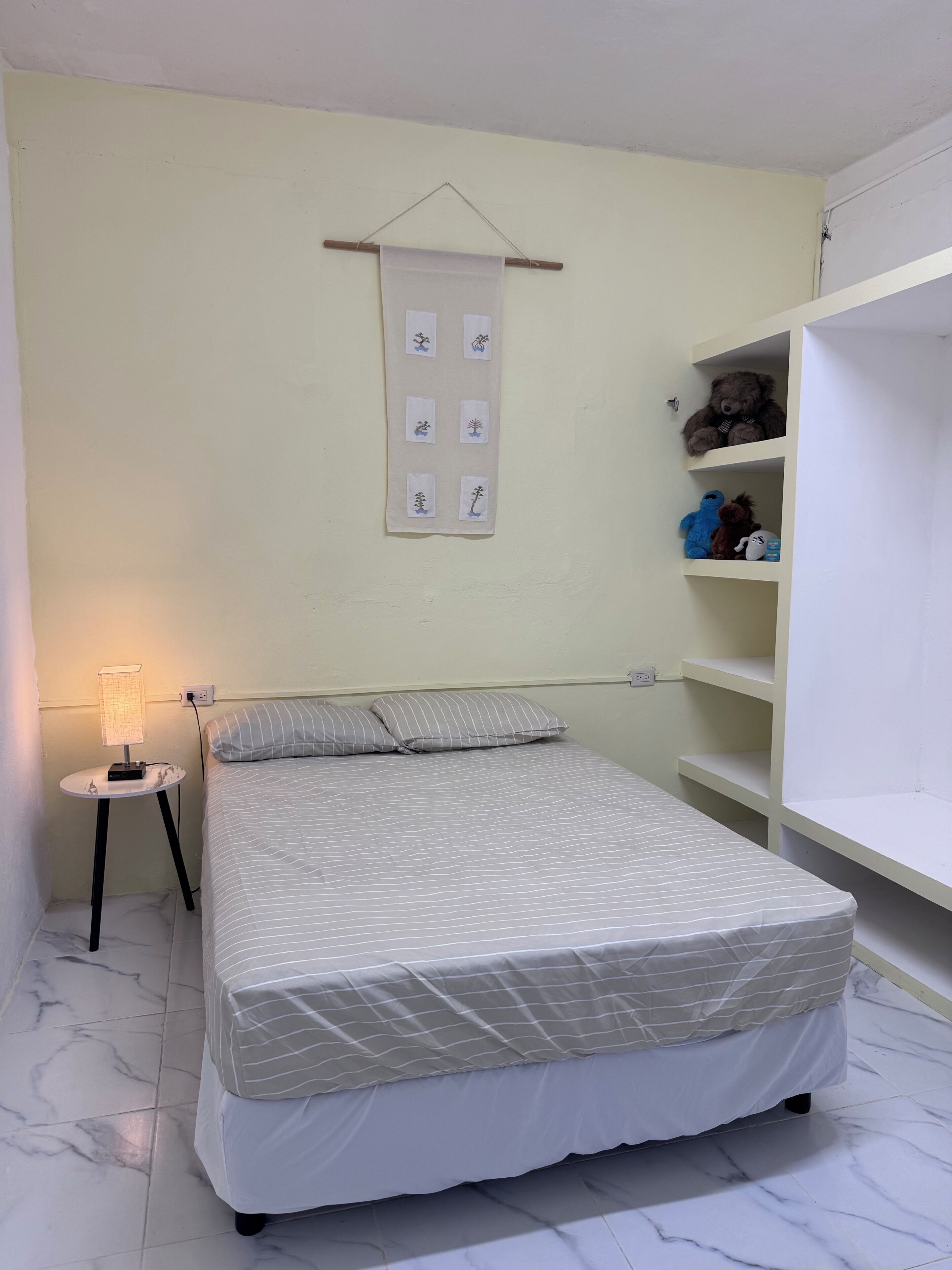 Een slaapkamer met een bed, een nachtkastje met een lamp, een wandplank met knuffels, en een wanddecoratie.