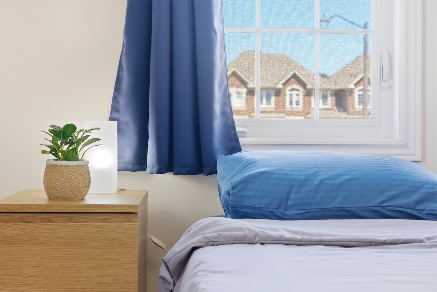 Une chambre à coucher avec un lit, une table de chevet ornée d’une plante, une fenêtre avec des rideaux et une vue sur les maisons à l’extérieur.