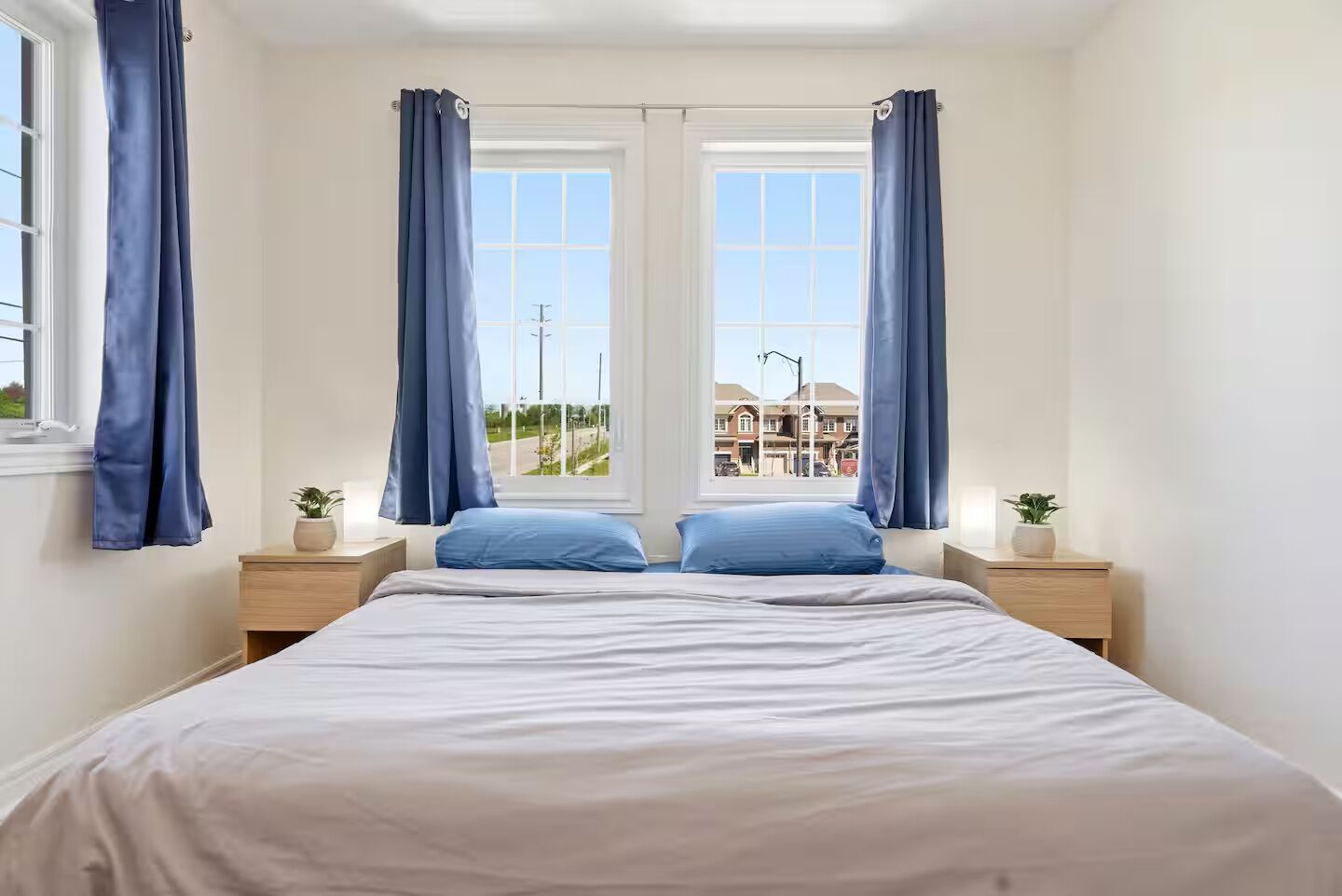 A bedroom with a bed, two nightstands, two potted plants, and a view of buildings through a window.