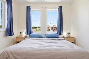 Une chambre à coucher avec un lit, deux tables de chevet, deux plantes en pot et une vue sur des immeubles par la fenêtre.