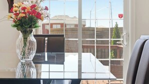 On aperçoit clairement un balcon avec une table et des chaises, un vase rempli de fleurs et une fenêtre donnant sur des immeubles.