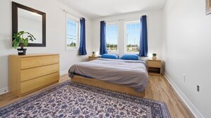 Une chambre à coucher comprenant un lit, une commode, une table de chevet, un miroir et une fenêtre avec des rideaux.