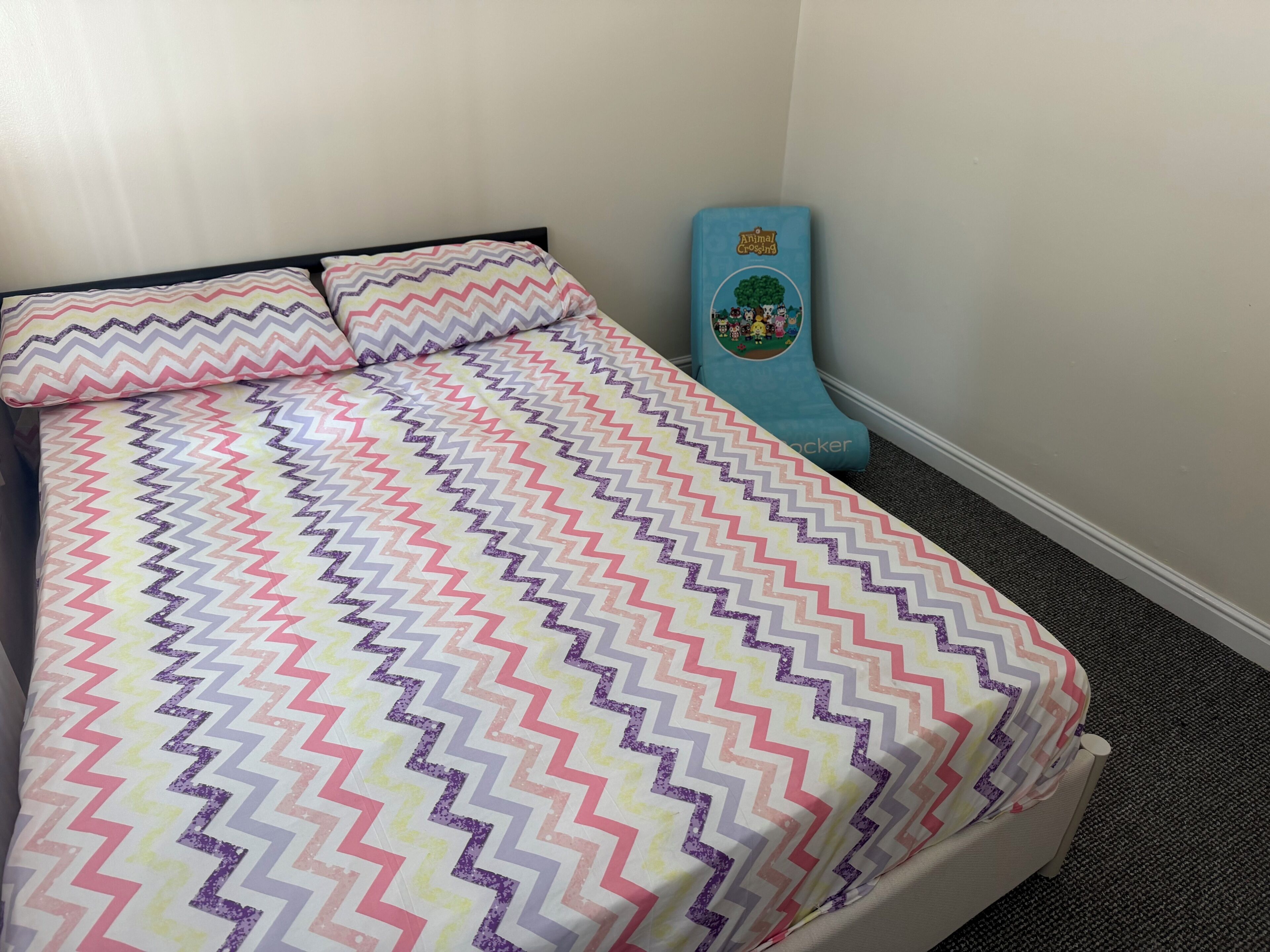 A bed with a patterned bedspread and pillows, a blue mattress topper with a cartoon design, and a carpeted floor.