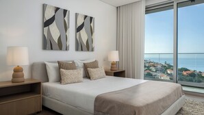 A modern hotel room with a large bed, two bedside tables with lamps, a view of the sea, and abstract wall art.