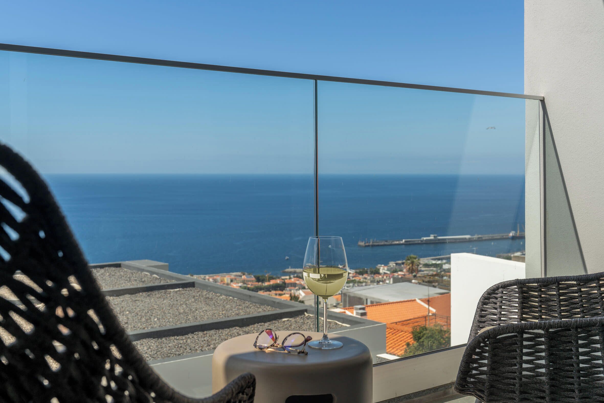 Ein Balkon mit Glastisch, auf dem ein Weinglas und zwei Brillen stehen, mit Blick auf die Küste, das Meer und eine Brücke.