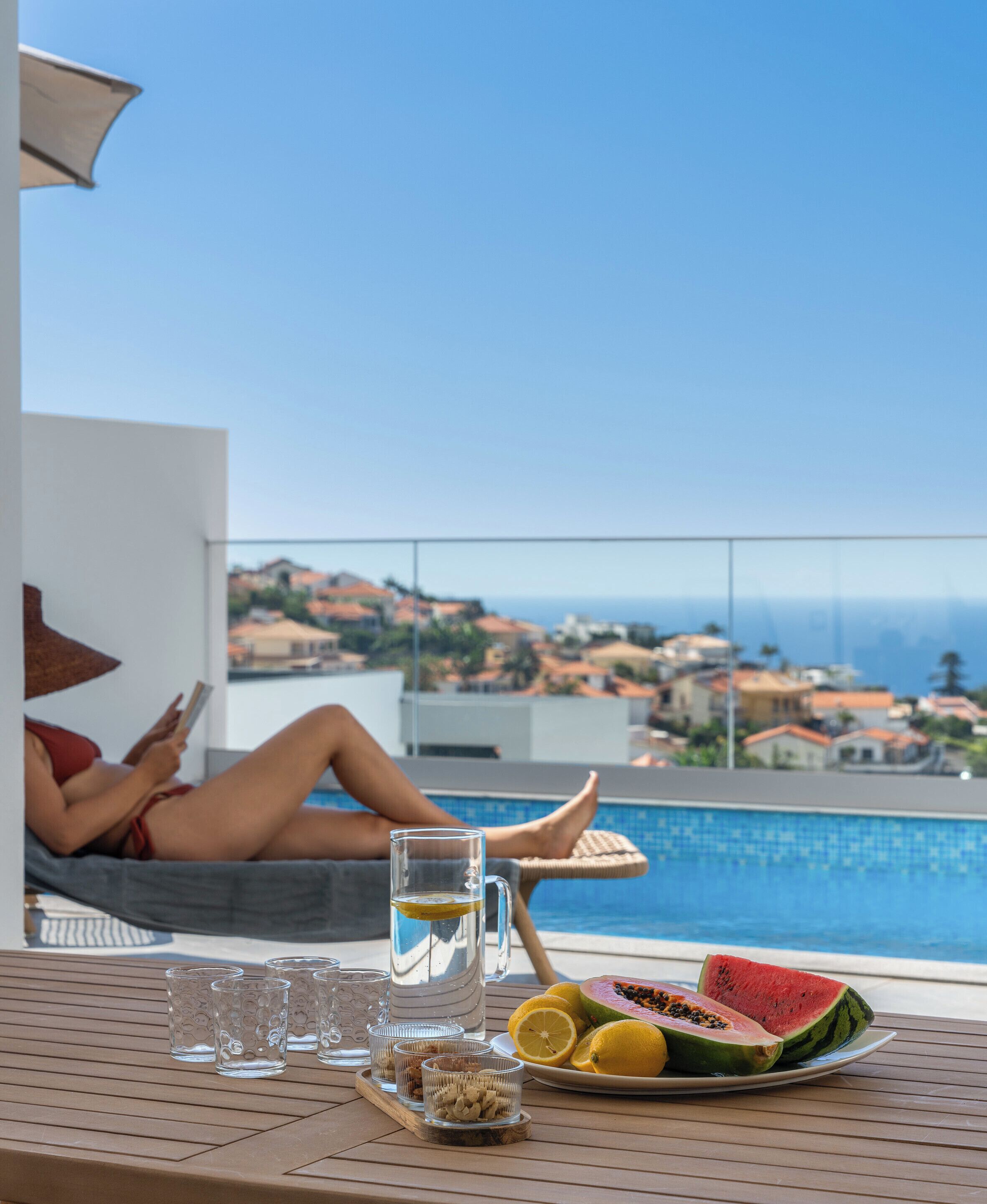 Eine Person entspannt am Pool mit Blick auf das Meer und die Hügel, ein Tisch mit Getränken und Obst, und ein klarer Himmel.