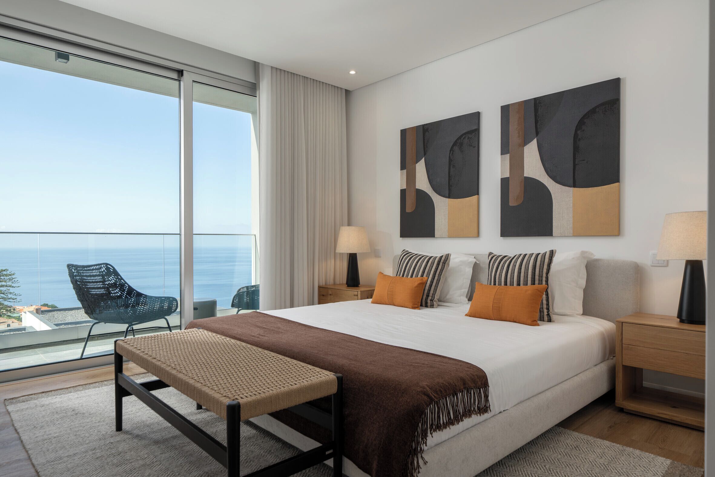 A modern hotel room with a large bed, a view of the sea, and abstract artwork on the wall.