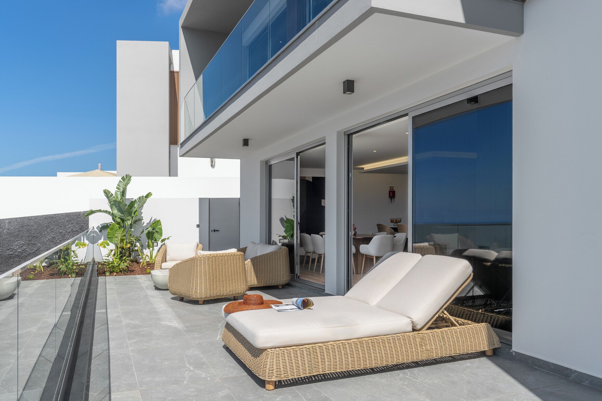 Eine moderne Lounge-Bereich im Freien mit Korbmöbeln, einer Glastür, die zu einem Innenraum führt, und Blick auf den Himmel.