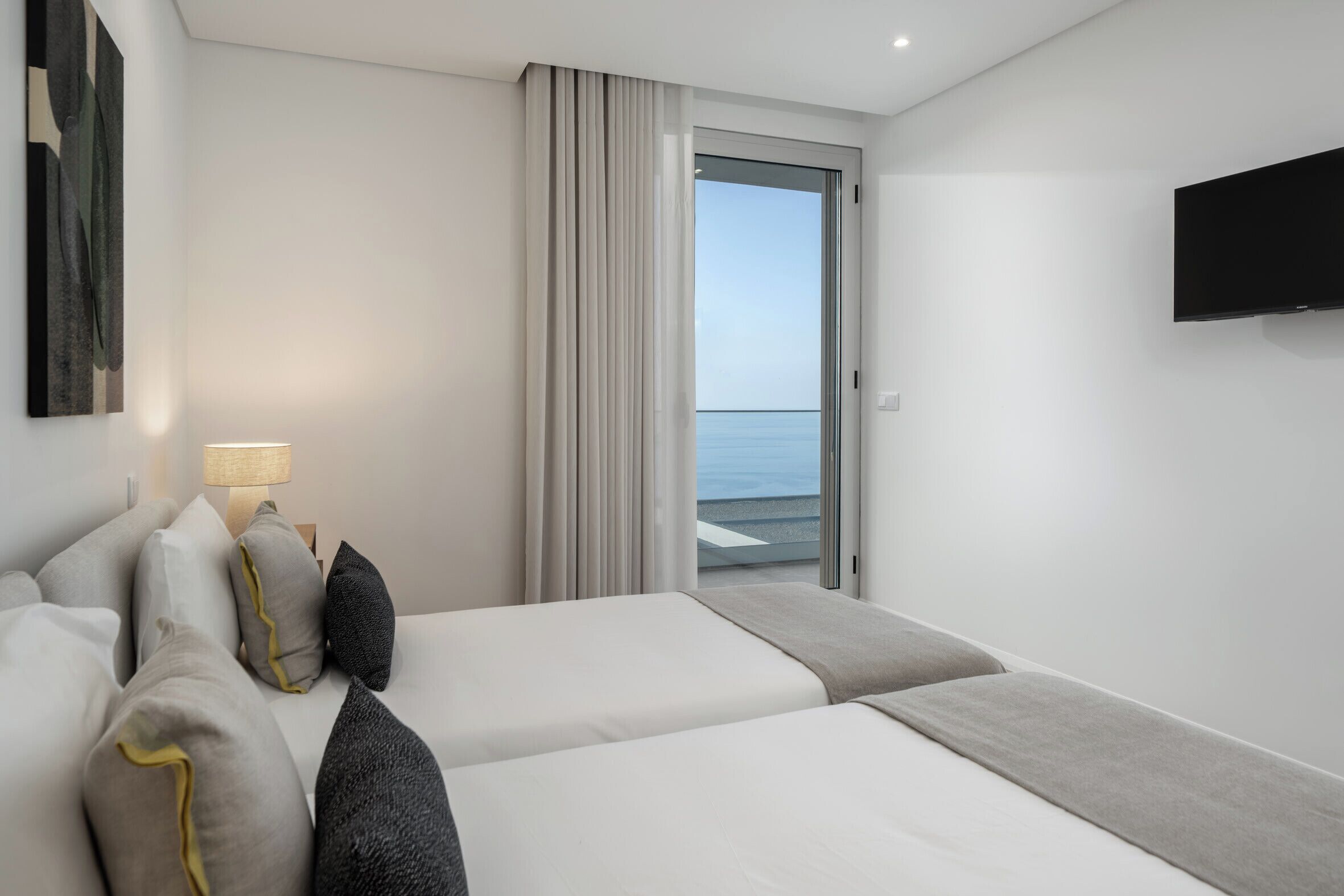 A hotel room with two beds, a TV, a lamp, and a view of the sea through a sliding door.