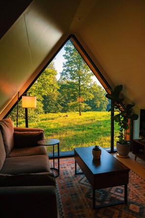 A cozy room with a sloped ceiling, a sofa, a small table, and a view of a green field through a large window.