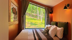 Ein Schlafzimmer mit einem großen Fenster, einem Bett mit gemusterten Betttextilien, einem Bild an der Wand und einer Lampe auf dem Nachttisch.