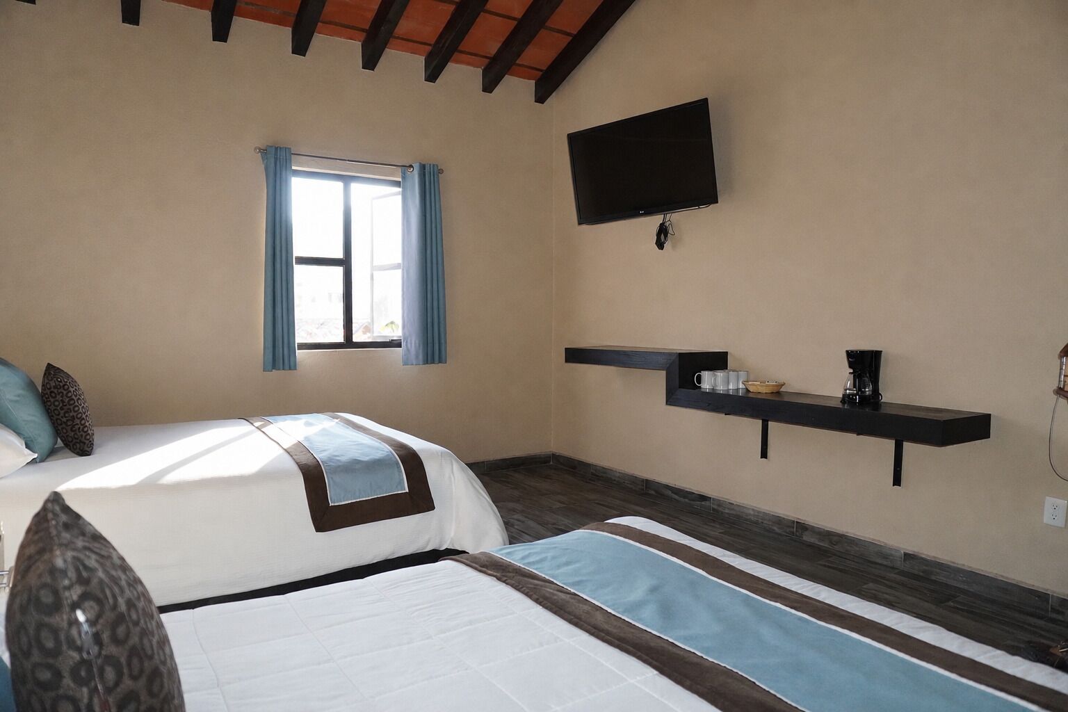 Une chambre d’hôtel avec un lit, une fenêtre avec des rideaux, un téléviseur fixé au mur et une étagère avec une machine à café.