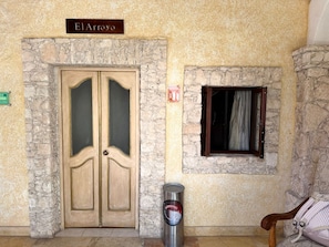 A stone wall entrance with a double wooden door, a small window with a curtain, and a trash can. - CABAÑAS BERNAL & HOTEL & SUITES (Ezequiel Montes)