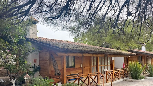 A wooden cabin with a tiled roof, surrounded by lush greenery and a stone-paved patio. - CABAÑAS BERNAL & HOTEL & SUITES (Quéretaro)
