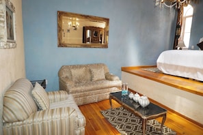 Una habitación de hotel con una cama, un sofá, un sillón estampado, una mesita con botellas y un espejo decorativo en la pared.