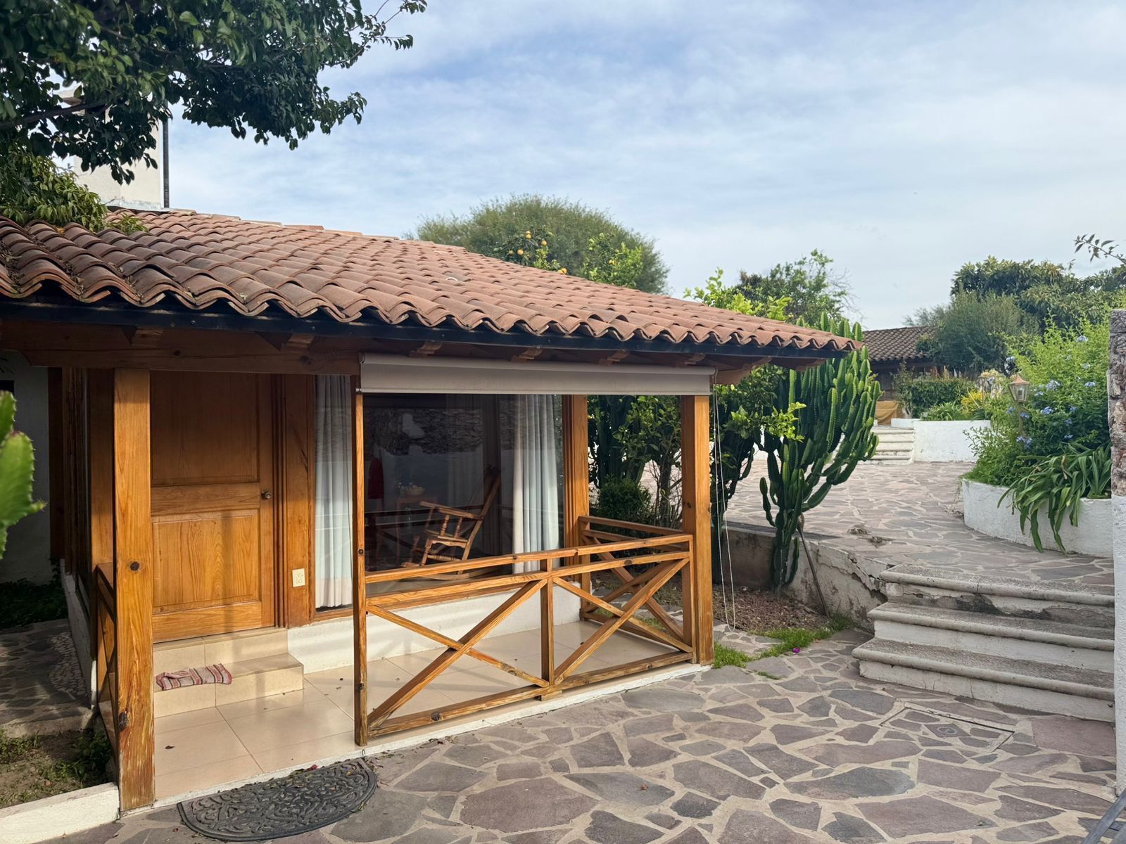 Una pequeña estructura de madera con techo de tejas, puerta de cristal y un banco de madera afuera.