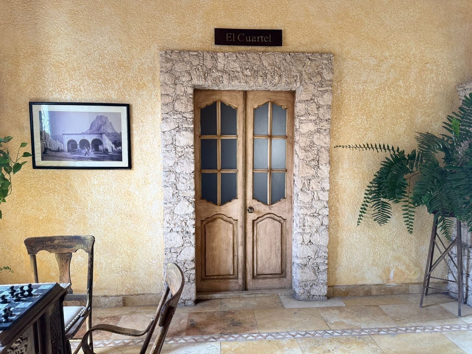A room with a stone wall, a wooden door labeled 'El Cuartel,' a framed photograph, a chess table, and a potted plant.