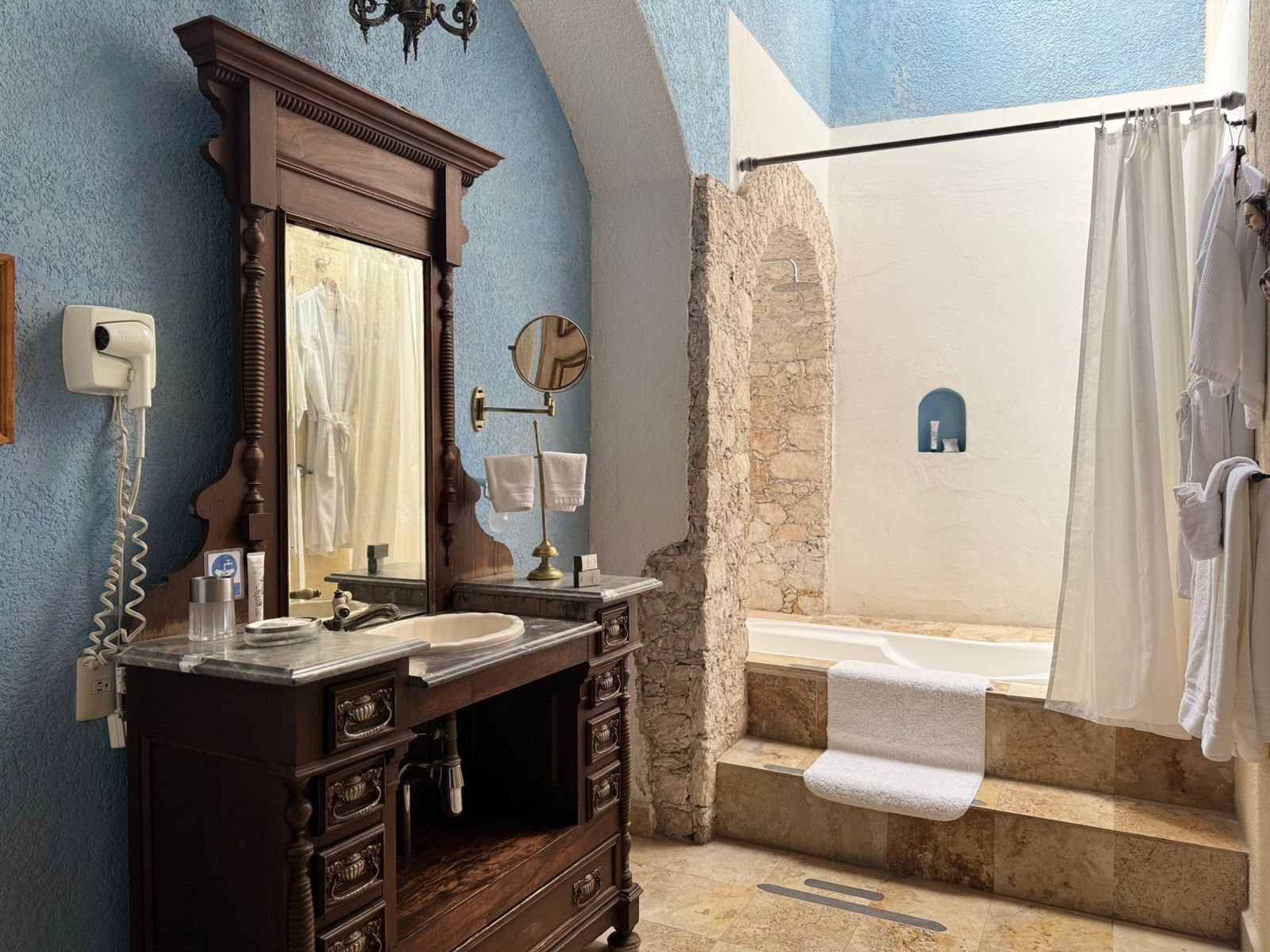A bathroom with a stone wall, a bathtub, a shower, a vanity with a mirror, and a towel rack.