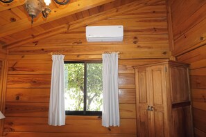 A wooden cabin interior with a window, white curtains, a white air conditioning unit, and a wooden cabinet. - CABAÑAS BERNAL & HOTEL & SUITES (Ezequiel Montes)