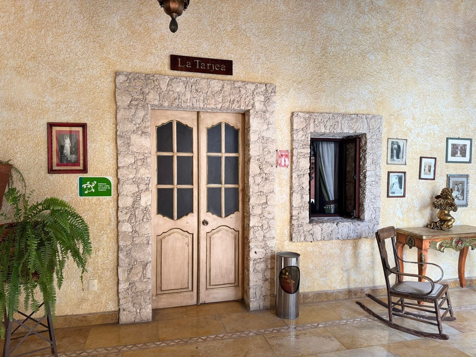 Una habitación con pared de piedra, una puerta doble con la leyenda 'La Tarjca', un ventanal pequeño con cortina, un cuadro enmarcado y una planta.