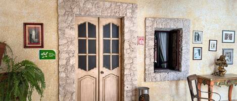 A room with a stone wall, a double door labeled 'La Tarjca,' a small window with a curtain, a framed picture, and a plant.