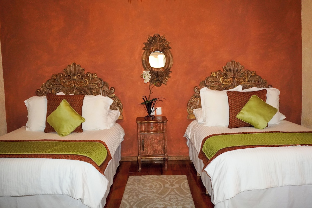 Una habitación de hotel con dos camas, cada una con un cabecero decorativo, una mesita de noche con una lámpara y un espejo en la pared.